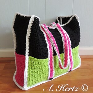 Op de afbeelding: Een zwarte, groene en roze gebreide boodschappentas met witte bies. De tas heeft twee lange hengsels en een knoopsluiting. De tekst "A. Hertz &copy; Crochet It Baby!" is zichtbaar onderaan de afbeelding.