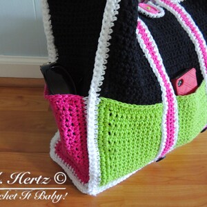 Op de afbeelding: Een zwarte, groene en roze gebreide tote bag met witte bies. De tas heeft twee zakken, &eacute;&eacute;n aan elke kant, en een witte band. De tas ligt op een houten oppervlak. "A. Hertz &copy; Crochet It Baby!" staat onderaan de afbeelding.