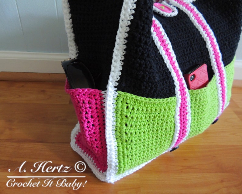 Op de afbeelding: Een zwarte, groene en roze gebreide tote bag met witte bies. De tas heeft twee zakken, &eacute;&eacute;n aan elke kant, en een witte band. De tas ligt op een houten oppervlak. "A. Hertz &copy; Crochet It Baby!" staat onderaan de afbeelding.