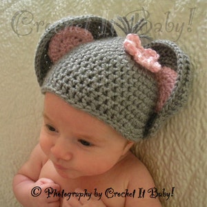 May include: A gray crocheted baby hat with pink ears and a pink bow. The hat is on a baby's head. The text "Photography by Crochet It Baby!" is at the bottom of the image.