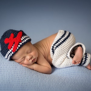 May include: A newborn baby wearing a blue and white striped crocheted hat with a red star and matching crocheted pants, sleeping on a blue blanket.