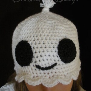 Op de afbeelding: Witte gehaakte geestenmuts met zwarte ogen en een gestikte glimlach. De muts heeft een witte pompon bovenop. "Crochet It Baby!" staat in zwarte tekst boven de muts.
