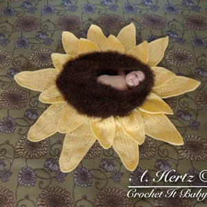 May include: A crocheted sunflower baby nest with a brown fuzzy center. A baby is lying in the nest. The nest is on a green floral fabric background. The text "L. Hertz &copy; Crochet It Baby!" is in the bottom right corner of the image.