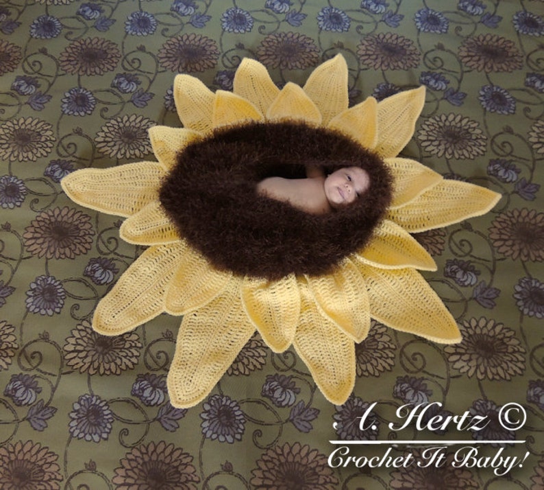 May include: A crocheted sunflower baby nest with a brown fuzzy center. A baby is lying in the nest. The nest is on a green floral fabric background. The text "L. Hertz &copy; Crochet It Baby!" is in the bottom right corner of the image.