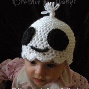Op de afbeelding: Een witte gehaakte baby muts met zwarte ogen en een glimlachende mond. De muts heeft een witte pompon bovenop. De muts wordt gedragen door een baby met een roze shirt.