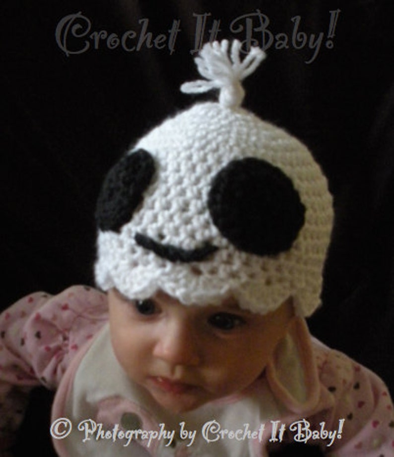 Op de afbeelding: Een witte gehaakte baby muts met zwarte ogen en een glimlachende mond. De muts heeft een witte pompon bovenop. De muts wordt gedragen door een baby met een roze shirt.