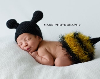 May include: A baby wearing a black crocheted bee costume with yellow and black stripes. The baby is sleeping on a white blanket.