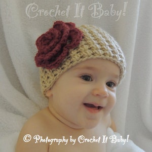 May include: A baby wearing a beige crocheted hat with a burgundy flower on top. The hat is crocheted with a textured stitch. The baby is smiling.