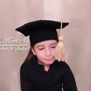 May include: A child wearing a black graduation cap with a gold tassel. The cap is crocheted and has a square top. The child is looking down and has a serious expression on their face. The text "A. Hertz © Crochet It Baby!" is visible in the background.