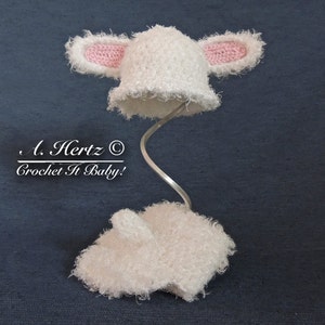 May include: White crocheted baby hat with pink ears and a fluffy texture. The hat is displayed on a metal stand. The text "A. Hertz © Crochet It Baby!" is visible below the hat.