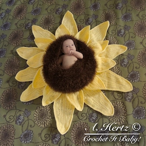 May include: A crocheted sunflower baby prop with a brown fuzzy center. A baby is laying in the center of the sunflower. The sunflower is on a green and brown floral patterned fabric.