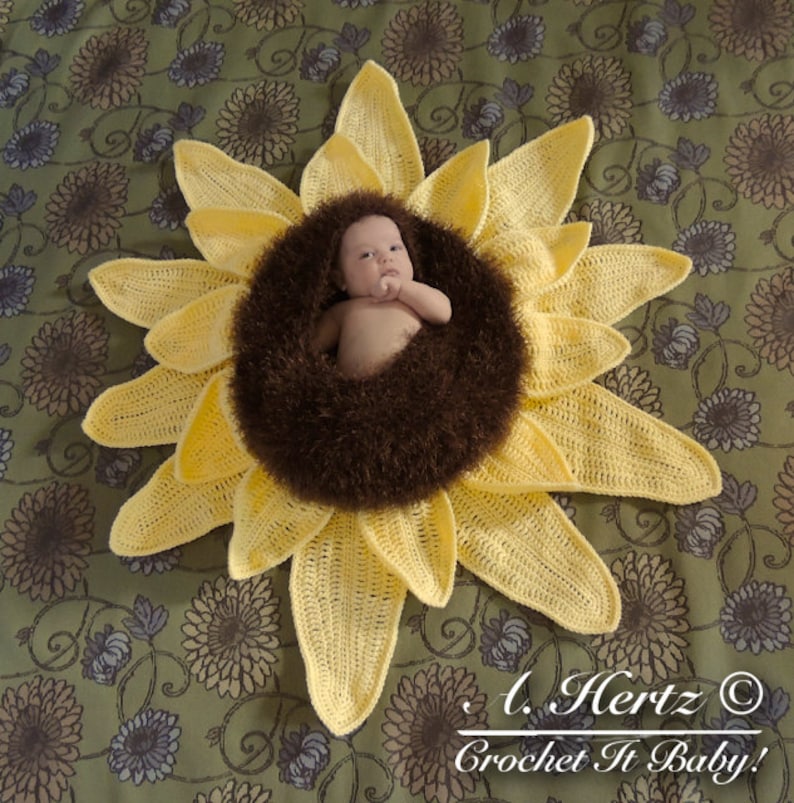 May include: A crocheted sunflower baby prop with a brown fuzzy center. A baby is laying in the center of the sunflower. The sunflower is on a green and brown floral patterned fabric.