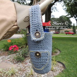 Op de afbeelding: Een gehaakte blauwe bekerhouder voor een kinderwagen met een zak en twee bronzen knopen. De houder is bevestigd aan een kinderwagenhandvat. De tekst "A. Hertz Crochet It Baby!" is zichtbaar op de achtergrond.
