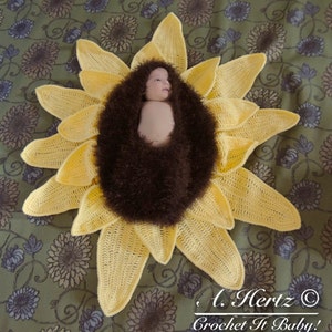 May include: A crocheted sunflower baby prop with a brown fuzzy center. A baby is lying inside the sunflower. The text "L. Hertz &copy; Crochet It Baby!" is at the bottom of the image.