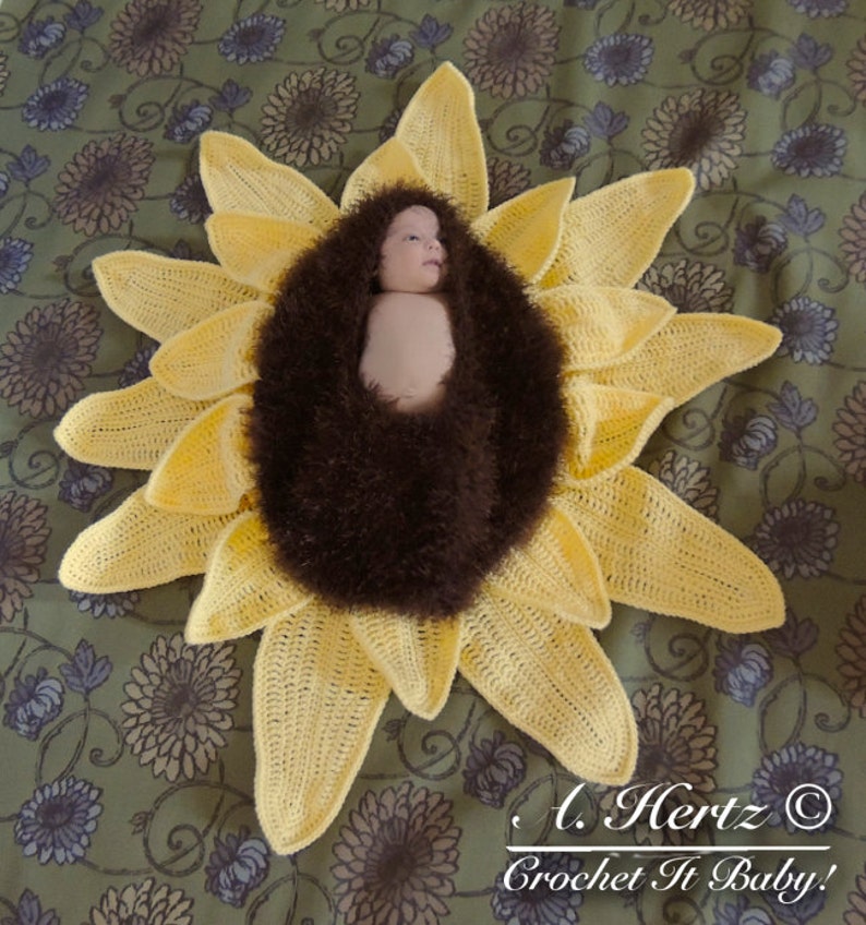 May include: A crocheted sunflower baby prop with a brown fuzzy center. A baby is lying inside the sunflower. The text "L. Hertz &copy; Crochet It Baby!" is at the bottom of the image.