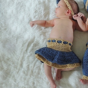 May include: A baby wearing a blue and tan crocheted skirt and headband. The headband has a bow on top. The baby is lying on a white fluffy surface.