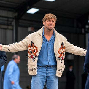 May include: A cream-colored chunky knit cardigan featuring a prominent fox face graphic on the front panels, a wide collar, and ribbed cuffs. The sweater is worn over a blue button-down shirt and light blue denim jeans.