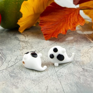 May include: Two small, white, ghost-shaped glass beads with smooth, glossy surfaces. Each bead features two black, circular eyes and a larger, black, oval-shaped mouth, creating a simple, expressive face on the front of the white glass body.