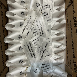 May include: A collection of white satin ribbons folded into awareness loops, each featuring a black printed diamond icon, the name Sheila Diana Miller, the years 1964-2023, and the text Forever In Our Hearts, always on our mind, secured with gold pins.