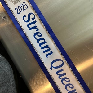 May include: A white sash with blue trim and the words "Stream Queen" in blue script. The sash also features the year "2025" and a silver crown graphic. Rhinestones line the edges of the sash.