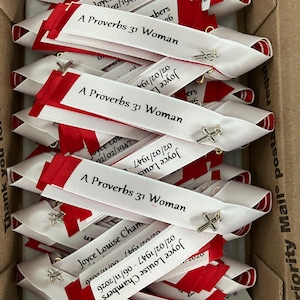 May include: A collection of white and red ribbon pins featuring a silver cross charm. Each ribbon is printed with the text A Proverbs 31 Woman and the name Joyce Louise Chambers with the dates 02/02/1947 - 06/11/2026 in black ink.