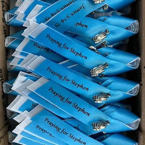 May include: A collection of light blue ribbon pins arranged in a box. Each ribbon features the printed text "Praying for Stephen" and "James 5:14-15" in black ink, accented with a small silver-toned metal charm shaped like a pair of praying hands.
