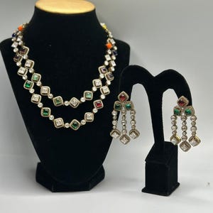 May include: A two-strand necklace and a pair of matching drop earrings featuring square-cut stones in red, green, and clear white, set in a metallic frame. The necklace has a layered design, and the earrings have a multi-tiered dangling structure.