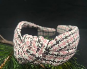 May include: A knotted headband with a pink, black, and white houndstooth pattern. The fabric-covered headband is a stylish hair accessory. The accessory is resting on a bed of green pine needles.