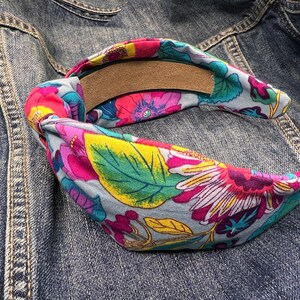 May include: A blue and gray fabric headband with a floral pattern in shades of pink, yellow, green, and white. The headband is tied in a knot at the back.