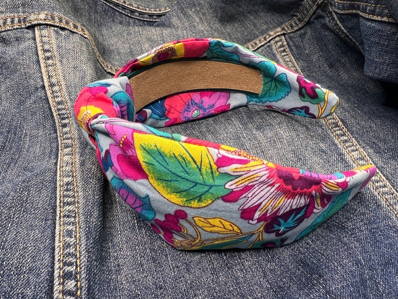May include: A blue and gray fabric headband with a floral pattern in shades of pink, yellow, green, and white. The headband is tied in a knot at the back.