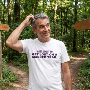 May include: A white short-sleeved crew neck t-shirt featuring the purple text "MOST LIKELY TO GET LOST ON A MARKED TRAIL" printed in a bold, sans-serif font across the center of the chest area, made of a standard cotton fabric.