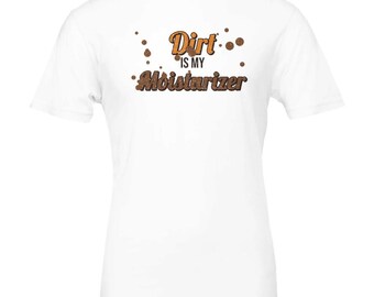Peut inclure: Un t-shirt blanc à manches courtes avec le texte Dirt IS MY Moisturizer en lettres marron et orange, accompagné de petits accents graphiques circulaires marron dispersés autour des mots sur le devant du vêtement.
