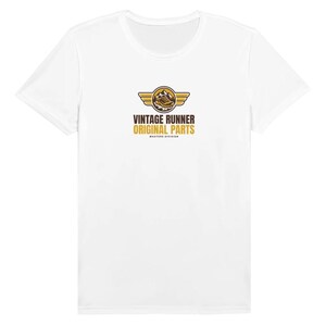 May include: A white short-sleeve t-shirt featuring a central graphic with a brown running shoe inside a circle with gold wings. Below the graphic, the text reads VINTAGE RUNNER ORIGINAL PARTS and MASTERS DIVISION in brown and gold.