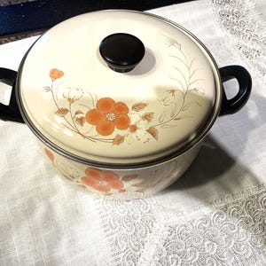May include: A cream-coloured enamel pot with a matching lid, featuring a floral pattern of orange and brown flowers. The pot has two black side handles and a black knob on the lid. The exterior surface is smooth and glossy with a rounded shape.