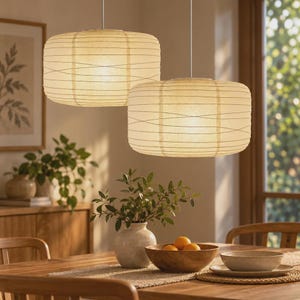 May include: Two cylindrical pendant lamps with off-white paper shades and thin horizontal wire ribbing. The lamps hang from the ceiling by thin white cords, emitting a warm, soft glow that illuminates the space below them.