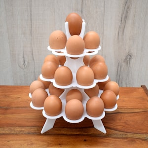 May include: A white three-tiered egg holder with 21 brown eggs. The holder is made of plastic and has a circular base with three legs.