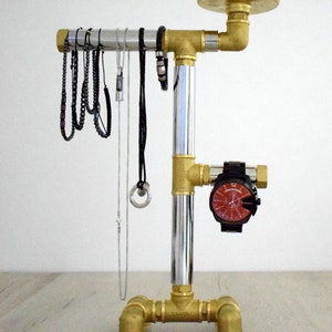 May include: A silver and brass jewelry stand with a round top and three legs. The stand has a silver pipe in the center and brass pipes on the sides. There are several necklaces and bracelets hanging from the stand. A watch is attached to the side of the stand.