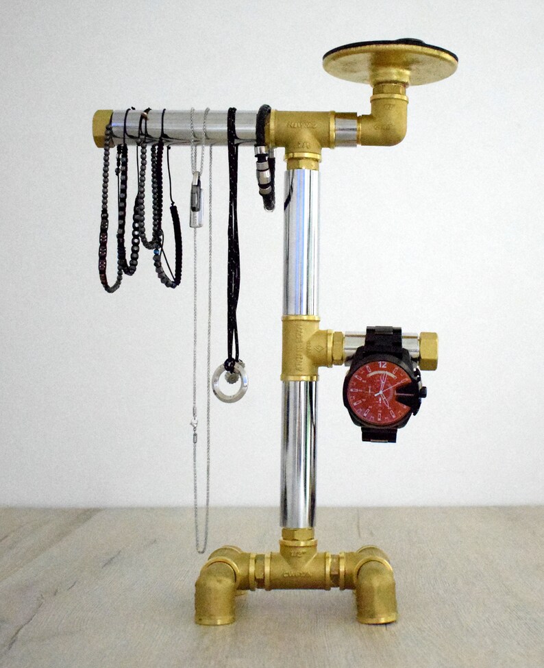 May include: A silver and brass jewelry stand with a round top and three legs. The stand has a silver pipe in the center and brass pipes on the sides. There are several necklaces and bracelets hanging from the stand. A watch is attached to the side of the stand.