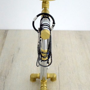 May include: A gold metal jewelry stand with a round top and four legs. The stand has a silver metal pipe in the center with multiple necklaces and bracelets hanging from it.