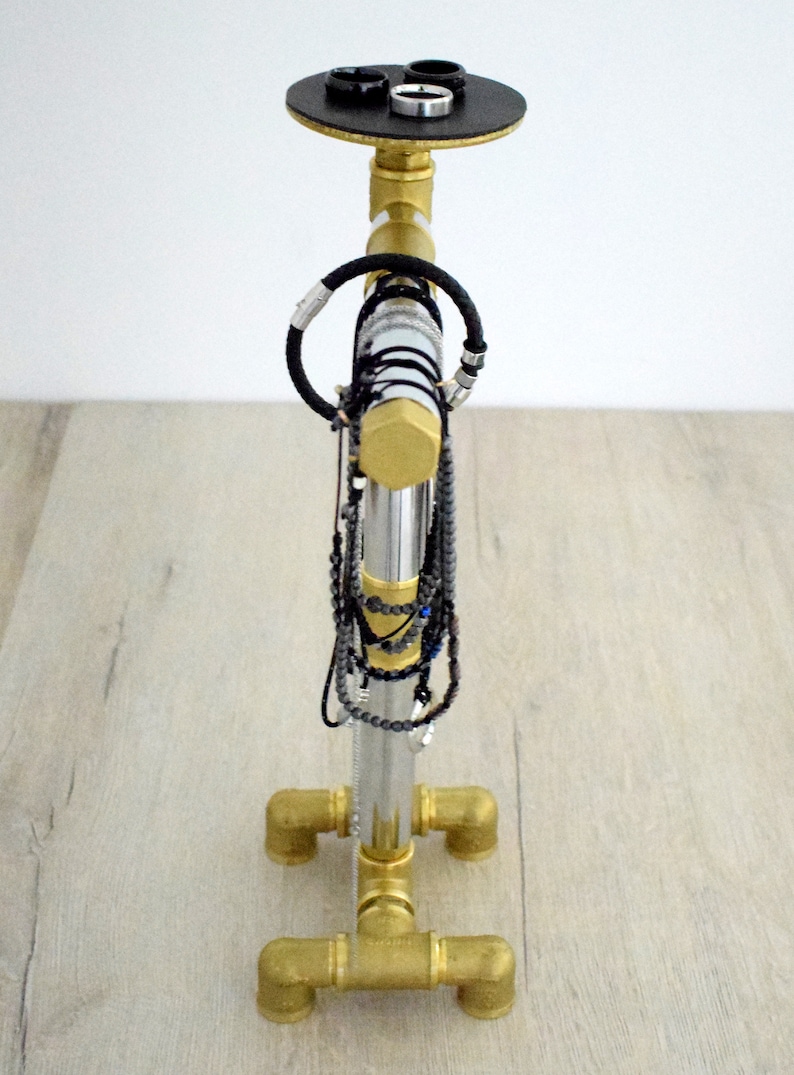 May include: A gold metal jewelry stand with a round top and four legs. The stand has a silver metal pipe in the center with multiple necklaces and bracelets hanging from it.