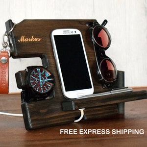 Può includere: Un supporto per telefono in legno marrone scuro con il nome personalizzato "Markus" inciso sul davanti. Il supporto ha uno spazio per un telefono, un orologio, una penna e occhiali.