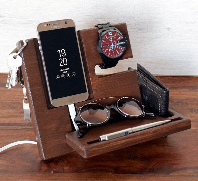 May include: A wooden organiser with compartments for a phone, watch, keys, sunglasses, and a pen. The phone is a Samsung Galaxy displaying the time as 19:20. The watch has a black strap and a red face with white markings. The organiser is made of dark brown wood.