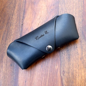 May include: Black leather glasses case with a silver button closure. The case is engraved with the name "Costas G."