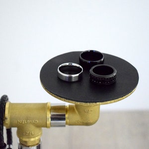 May include: Three rings on a black circular platform. The platform is attached to a gold pipe fitting. One ring is silver, and the other two are black.