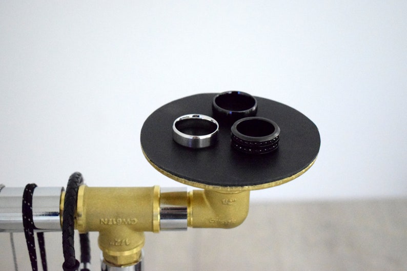 May include: Three rings on a black circular platform. The platform is attached to a gold pipe fitting. One ring is silver, and the other two are black.