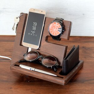 May include: A wooden organiser with slots for a phone, watch, glasses, and a pen. The phone is a black Samsung Galaxy with the time 19:18 displayed on the screen. A brown leather wallet is in the bottom slot.
