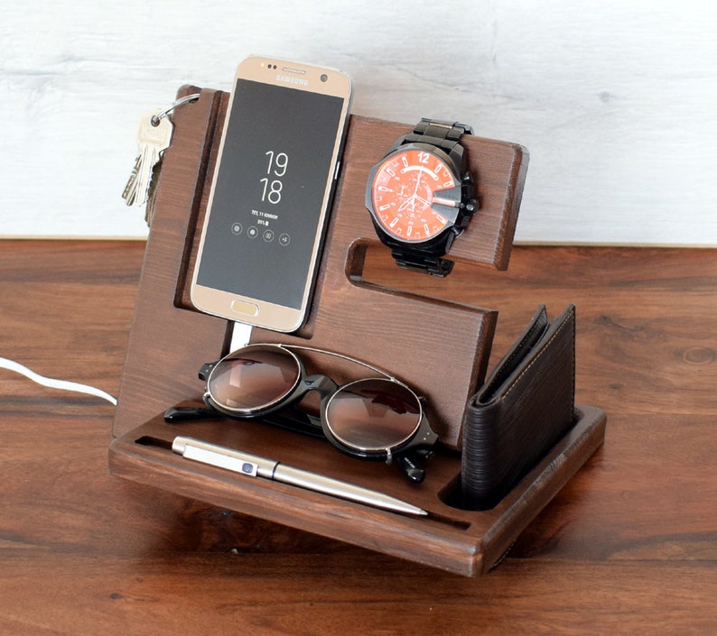 May include: A wooden organiser with slots for a phone, watch, glasses, and a pen. The phone is a black Samsung Galaxy with the time 19:18 displayed on the screen. A brown leather wallet is in the bottom slot.