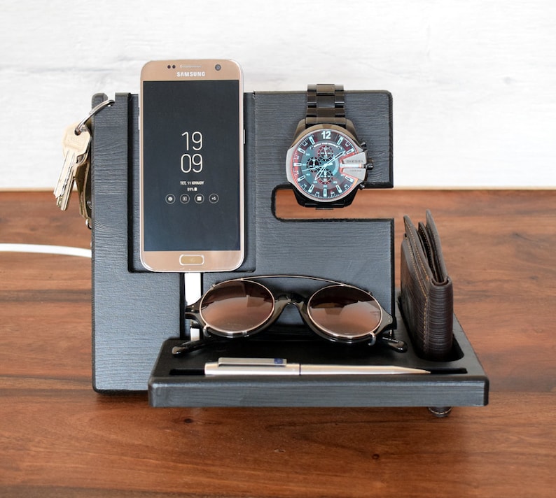 Puede incluir: Organizador de madera negro con ranuras para un tel&eacute;fono, un reloj, gafas de sol y una cartera. El tel&eacute;fono es un Samsung Galaxy con la hora 19:09 mostrada en la pantalla.