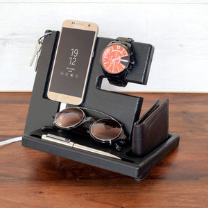 Puede incluir: Organizador de madera negro con ranuras para un tel&eacute;fono, un reloj, gafas de sol y un bol&iacute;grafo. El tel&eacute;fono es un Samsung Galaxy que muestra la hora 19:12. Tambi&eacute;n se incluye una cartera de cuero marr&oacute;n.