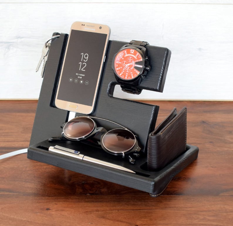 Puede incluir: Organizador de madera negro con ranuras para un tel&eacute;fono, un reloj, gafas de sol y un bol&iacute;grafo. El tel&eacute;fono es un Samsung Galaxy que muestra la hora 19:12. Tambi&eacute;n se incluye una cartera de cuero marr&oacute;n.